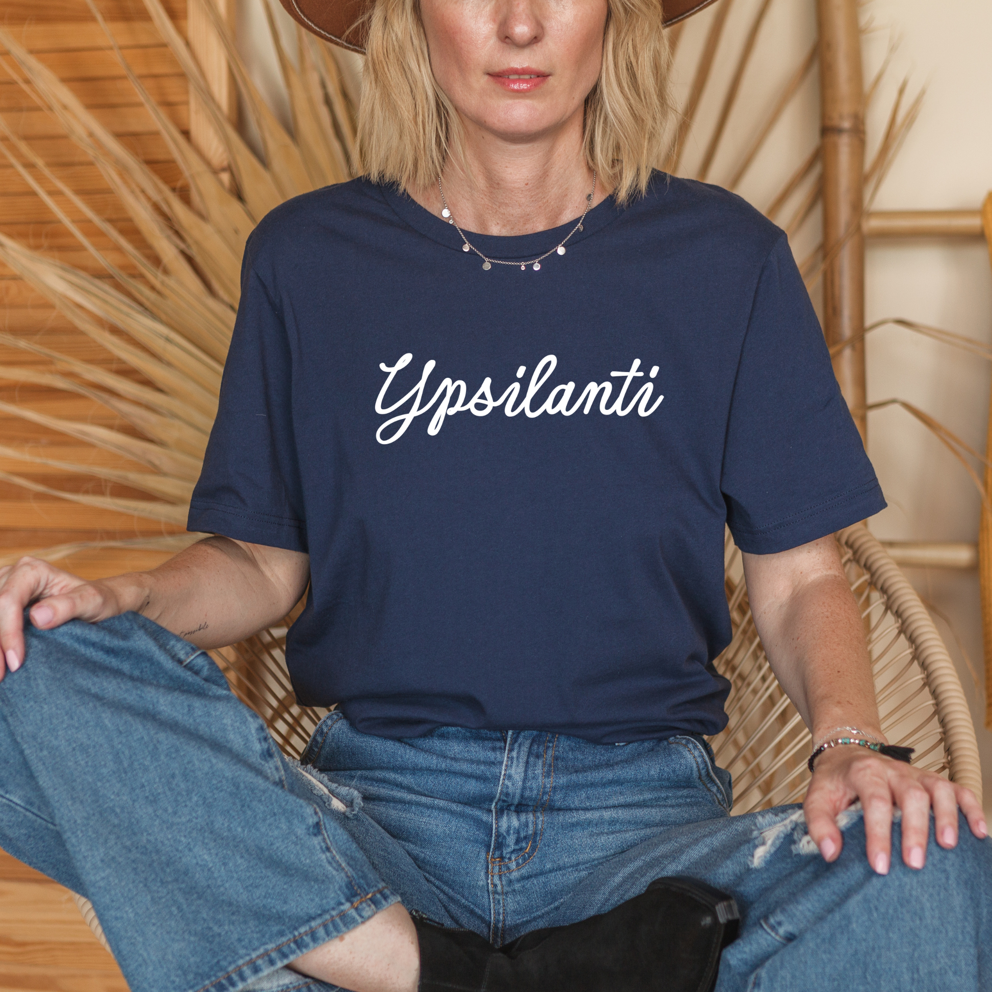 Person wearing a navy blue t-shirt with 'Ypsilanti' text, sitting on a woven chair.